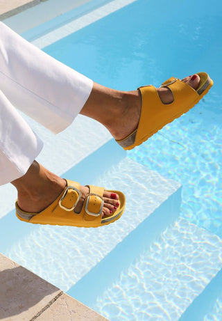 Gelbe Pantoletten aus Softnappaleder, getragen von einer Person am Pool. Die Schuhe haben zwei Schnallen.